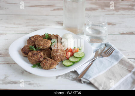 Saftige köstliche Fleisch Schnitzel in eine weiße Platte auf hellem Holztisch. Metballs gebraten serviert mit Tomaten und Gurken und Sauerrahm. Stockfoto