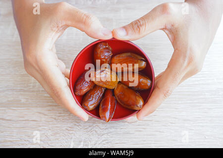 Hand, die rote Schale mit getrockneten Früchten Datum von Palm auf hölzernen Hintergrund, Phoenix dactylifera auf rot Schüssel in der Hand. Stockfoto