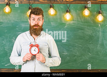 Lehrer in Brillen hält Wecker. Zeitplan und Regime Konzept. Mann mit Bart und Schnurrbart auf überrascht Gesicht im Klassenzimmer. Bärtige hipster hält, schwarzen Brettes auf Hintergrund, kopieren. Stockfoto