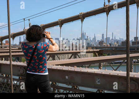 New York Tourist, Ansicht von hinten im Sommer eine alleinreisende weibliche Touristen stehen auf der Brooklyn Bridge und nehmen ein Foto von Midtown Manhattan, New York City, USA Stockfoto