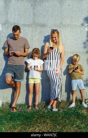 Fotos von der blonden Frau und Mann und Kinder mit Handys in den Händen stehend auf Betonwand auf der Straße im Sommer Tag Stockfoto