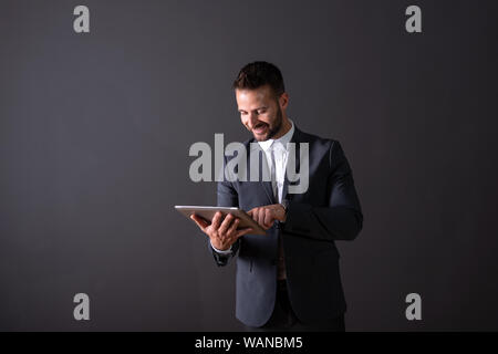 Ein lächelndes hübscher junger Geschäftsmann im Anzug mit einem Tablett vor einem dunklen Hintergrund grau in einem Studio. Stockfoto