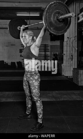 Eine hübsche blonde Frau mittleren Alters mit Sixpack ist ein Overhead sauber mit der Langhantel. Funktionelle Fitness und Kraftsport Training in einem Fitnessstudio. Stockfoto