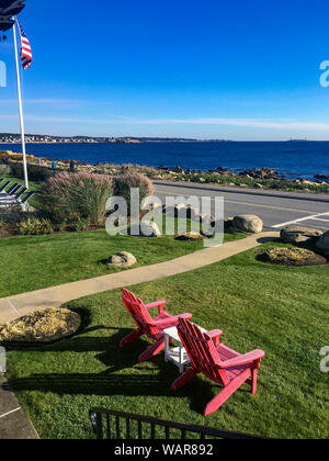 Gloucester, MA/USA, 19. Okt. 2018: Zwei rote Adirondack Stühle sitzen auf der Wiese mit Blick auf den Atlantik an einem schönen Herbsttag. Stockfoto