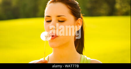 Junge Frau bläst Löwenzahn im Frühling. Blick von Außen. Freiheit und Spaß. Stockfoto