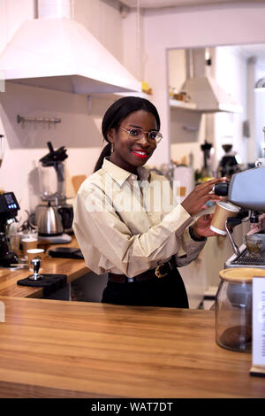 Lächelnd attraktive afrikanischen barista Vorbereitung eine frische Tasse Kaffee, während Sie sich an einer Kaffeemaschine im Cafe. Stockfoto