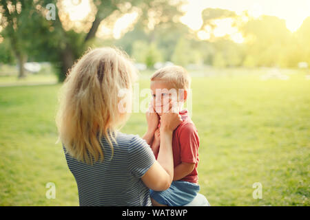 Jungen weißen Mutter ziehen junge Kleinkind Wangen. Mama sitzt zusammen mit Sohn auf Sommertag im Park und zeigt Ihre Liebe und Zuneigung. Happy Famil Stockfoto