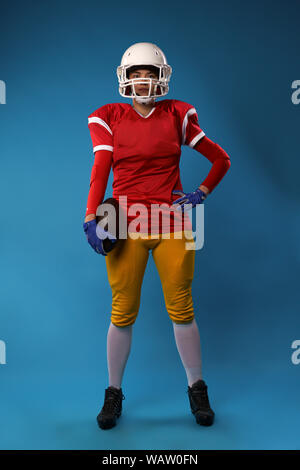 In voller Länge Foto der jungen amerikanischen Frau Fußballspieler in weißen Helm, Sportswear mit Ball in der Hand auf leeren blauen Hintergrund isoliert Stockfoto