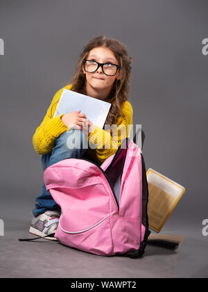 Spaß nette Schüler kind Mädchen in Brillen Ausblenden der Übungen Bücher mit schlechten Noten in der Nähe der Rucksack sitzt und schaut auf grauem Hintergrund. Closeup po Stockfoto