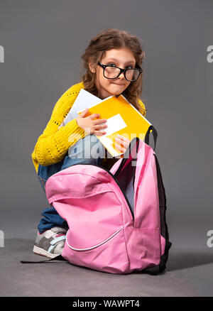 Spaß nette Schüler kind Mädchen in Brillen Ausblenden der Übungen Bücher mit schlechten Noten in der Nähe der Rucksack sitzt auf grauen Hintergrund. Closeup Portrait Stockfoto