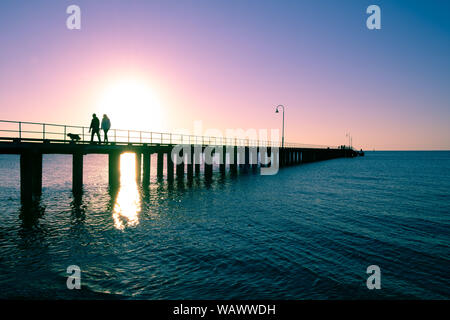 Paar mit Hund Silhouetten auf hölzernen Pier bei Sonnenuntergang Stockfoto