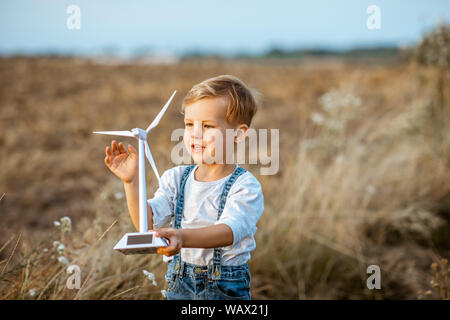 Neugierige junge Junge spielt mit Spielzeug Windenergieanlage im Feld, die studieren, wie grüne Energie funktioniert von einem jungen Alter Stockfoto