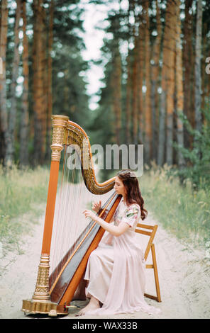 Frau Harfenistin sitzt am Waldweg und spielt Harfe im schönen Kleid vor dem Hintergrund der Kiefern. Stockfoto