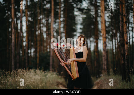 Frau harfenistin Spaziergänge im Wald und spielt Harfe im schönen Kleid vor dem Hintergrund der Kiefern. Stockfoto