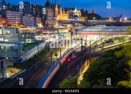 Blick von oben auf die Außenseite des Edinburgh Bahnhof mit einem Zug, der Plattform in der Nacht. Altstadt von Edinburgh Skyline im Hintergrund. Stockfoto