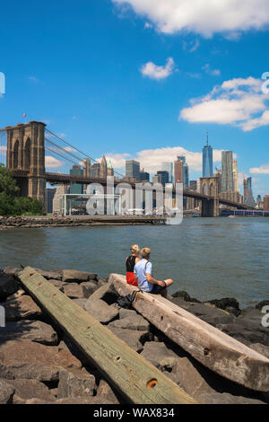 New York Paar, mit Blick auf die Brooklyn Bridge und Lower Manhattan Skyline von Pebble Beach Park mit zwei junge Menschen im Vordergrund, USA. Stockfoto