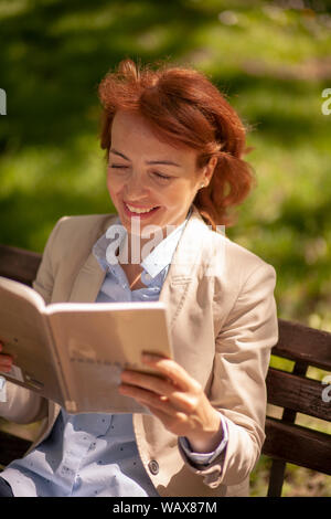 Porträt der lächelnde Frau, ein Buch lesen, in einem Park auf einer Bank sitzen. Stockfoto