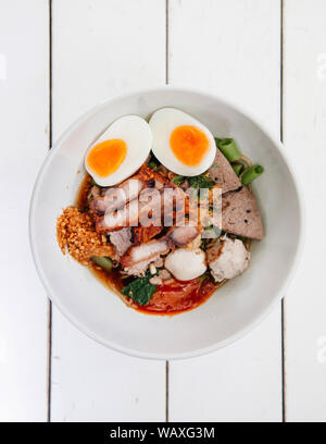 Thai Nudelsuppe mit Schweinefleisch, gekochte Eier, Leber, Fisch, Kugel, knuspriger Schweinebauch mit roten pikanter Sauce in weiße Schüssel. Ansicht von oben geschossen Stockfoto