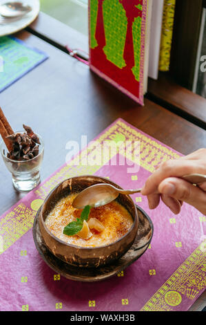 Französische Dessert, Löffel Graben in schöne Cremige Vanille Creme Brulee in Coconut Shell auf bunte Platte mat. Von der Seite Top Shot Stockfoto