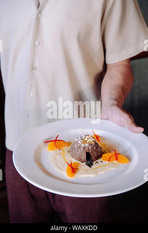 Französische Dessert. Schokolade Fondant lava Kuchen mit Vanillesauce, Zitrusfrüchte und Blattgold auf weiße Platte in der Kellner hand Stockfoto