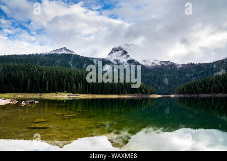 Montenegro, die ruhigen Gewässer des Schwarzen See im Nationalpark Durmitor von grünen Wald und hohe Schnee bedeckte Berge teilweise ausgeblendet Stockfoto