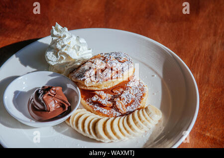 Fluffy pancake mit gebackenen Mandeln Schokolade Sauce die Sahne und Banane in weiße Platte auf Holz Tabelle unter Sonneneinstrahlung. Stockfoto