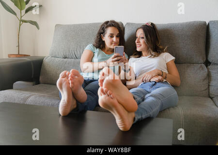 Zwei Mädchen in einer Couch mit Füßen in einer Tabelle mit einem Mobiltelefon Stockfoto