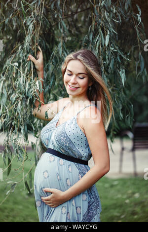 Glückliche gesunde Schwangerschaft und Mutterschaft. Portrait von Schwangeren jungen kaukasischen Frau mit langen blauen Kleid in Park außerhalb posieren. Schöne nachdenkliche Fe Stockfoto