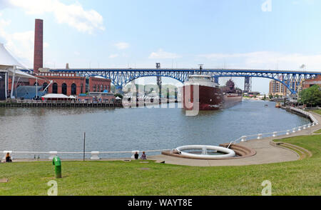 Ein Paar sitzt am Ufer der Flats East Bank entlang des Cuyahoga River, während ein Frachter im Sommer in Cleveland, Ohio, USA vorbeifährt. Stockfoto