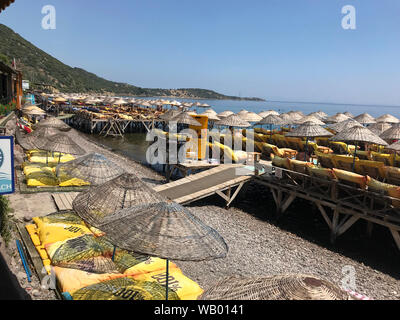 Direkt am Strand mit Liegestühlen und Sonnenschirmen auf Pfeilern über dem Wasser am Hafen von Assos an der Ägäischen Küste der Türkei. Stockfoto