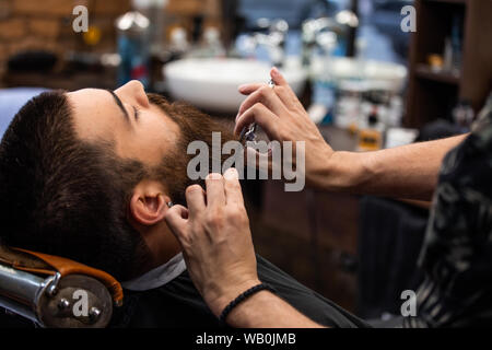 Close up 7/8-Foto von einem Friseur Arbeit für einen attraktiven Mann an der Friseur. Er tut Styling von seinen Bart mit der Schere Stockfoto