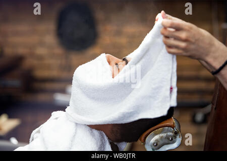 Friseur deckt das Gesicht eines Mannes mit einem warmen Handtuch. Traditionelle Ritual der Rasur des Bartes heiße und kalte Kompressen in einem alten Stil Friseur. Cli Stockfoto