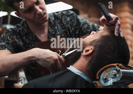 Close up 7/8-Foto von einem Friseur Arbeit für einen attraktiven Mann an der Friseur. Er tut Styling von seinen Bart mit der Schere Stockfoto