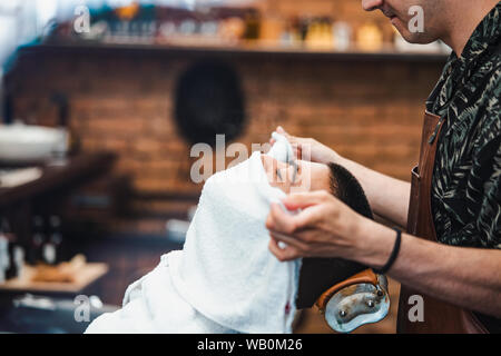 Friseur deckt das Gesicht eines Mannes mit einem warmen Handtuch. Traditionelle Ritual der Rasur des Bartes heiße und kalte Kompressen in einem alten Stil Friseur. Cli Stockfoto