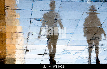 Unscharfe Reflexion Schatten Silhouetten von zwei Menschen zu Fuß auf einer nassen Straße an einem Sommertag in der Stadt Stockfoto