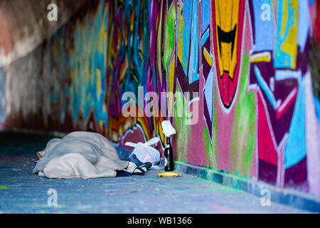 Mannheim, Deutschland. 23 Aug, 2019. In eine Decke gehüllt, einen Obdachlosen schläft in einer Brücke Durchfahrt auf eine Wand mit Graffiti bemalt. Neben ihm ist eine Flasche Bier und eine Banane ist auf dem Boden liegt. Foto: Uwe Anspach/dpa/Alamy leben Nachrichten Stockfoto