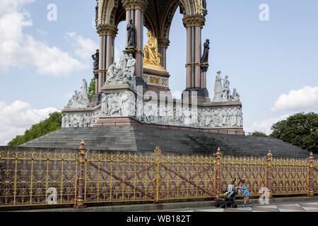 Ein Kind schiebt einen Kinderwagen vor der Golden Schmiedearbeiten an der Albert Memorial in Kensington Park, am 20. August 2019, in London, England. Das Albert Memorial, direkt nördlich der Royal Albert Hall in Kensington Gardens, London, wurde von Königin Victoria zum Gedenken an ihren geliebten Ehemann Prinz Albert, der im Jahr 1861 in Betrieb genommen. Von Sir George Gilbert Scott im Neugotischen Stil gestaltet, es hat die Form eines verzierten Baldachin oder Pavillon 176 Fuß hoch, im Stil einer gotischen Ziborium über dem Hochaltar der Kirche beherbergt eine Statue der Prinz auf der Südseite. Es dauerte mehr als zehn Jahre Stockfoto