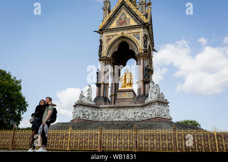 Ein paar Hug vor der Golden Schmiedearbeiten an der Albert Memorial in Kensington Park, am 20. August 2019, in London, England. Das Albert Memorial, direkt nördlich der Royal Albert Hall in Kensington Gardens, London, wurde von Königin Victoria zum Gedenken an ihren geliebten Ehemann Prinz Albert, der im Jahr 1861 in Betrieb genommen. Von Sir George Gilbert Scott im Neugotischen Stil gestaltet, es hat die Form eines verzierten Baldachin oder Pavillon 176 Fuß hoch, im Stil einer gotischen Ziborium über dem Hochaltar der Kirche beherbergt eine Statue der Prinz auf der Südseite. Es dauerte mehr als zehn Jahre zu Ergänzungen Stockfoto