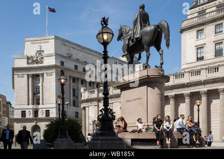 Mittag Stadt Arbeitnehmer genießen Sie warmen Sommer Sonnenschein unter dem Herzog von Wellington's Statue, steht gegenüber der Bank von England (rechts) bei der Bank Dreieck in der Stadt London, der Bezirk der Hauptstadt (aka der Square Mile), am 22. August 2019, in London, England. Stockfoto