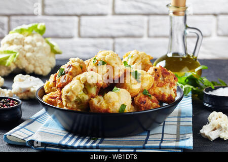 Blumenkohlröschen gebraten in Teig in eine Schüssel auf einen konkreten Tisch mit frischen Blumenkohl und eine Flasche Olivenöl serviert auf dem Hintergrund einer Brick w Stockfoto