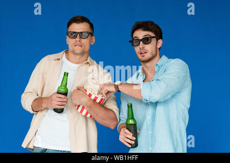 Zwei Freunde in einer 3D-Brille holding Flaschen Bier und Popcorn isoliert auf Blau Stockfoto