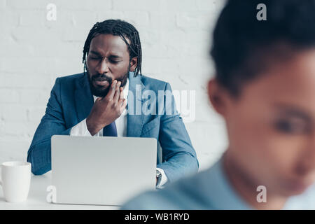 Unglücklich afrikanische amerikanische Geschäftsmann Leiden von Zahn Schmerzen beim Sitzen im Büro in der Nähe der hübschen Kollegen Stockfoto