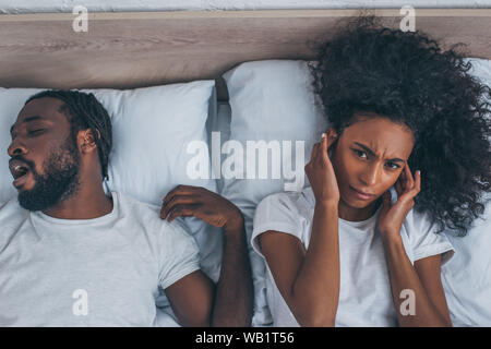 Blick von oben auf die afrikanische amerikanische Frau leidet unter Kopfschmerzen, während im Bett liegend in der Nähe von Schnarchen Mann Stockfoto