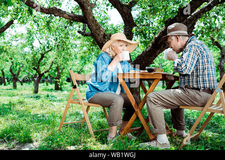 Ältere Frau und Mann sitzen auf Stühlen und trinken Kaffee in der Nähe von Bäumen Stockfoto