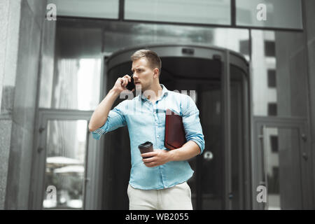 Junge schöne business Mann mit Tasse Kaffee und Laptop in den Händen der Unterhaltung am Telefon steht in der Nähe der Eingang zum Büro business center Stockfoto