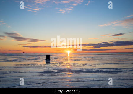 Die Sonne über einen gefrorenen Hafen und ein Eis angeln shack in ländlichen Prince Edward Island, Kanada. Leuchtturm am Horizont. Stockfoto