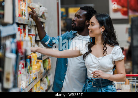 Selektiver Fokus der stattlichen african american zeigte mit dem Finger in der Nähe von attraktiven asiatischen Frau auf der Suche nach Regale mit Lebensmitteln Stockfoto