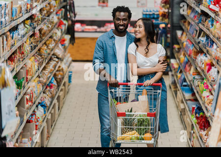 Selektiver Fokus der asiatischen Frau glücklich und afrikanische amerikanische Mann mit Einkaufswagen im Supermarkt Stockfoto