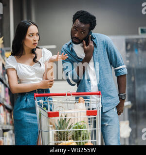 Verärgert asiatische Frau gestikuliert während Sie afrikanische amerikanische Mann auf dem Smartphone im Supermarkt Stockfoto