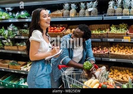 Gerne afrikanische amerikanische Mann an asiatische Frau suchen mit Notebook in der Nähe der Warenkorb mit Lebensmitteln Stockfoto